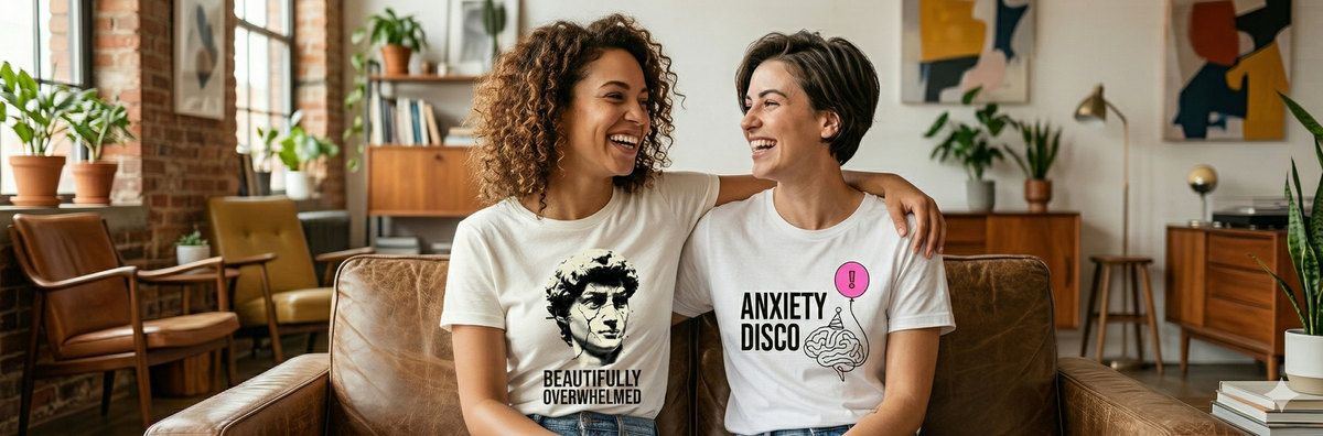 Two women sitting on a couch laughing with each other, wearing humorous anxiety T-shirts.